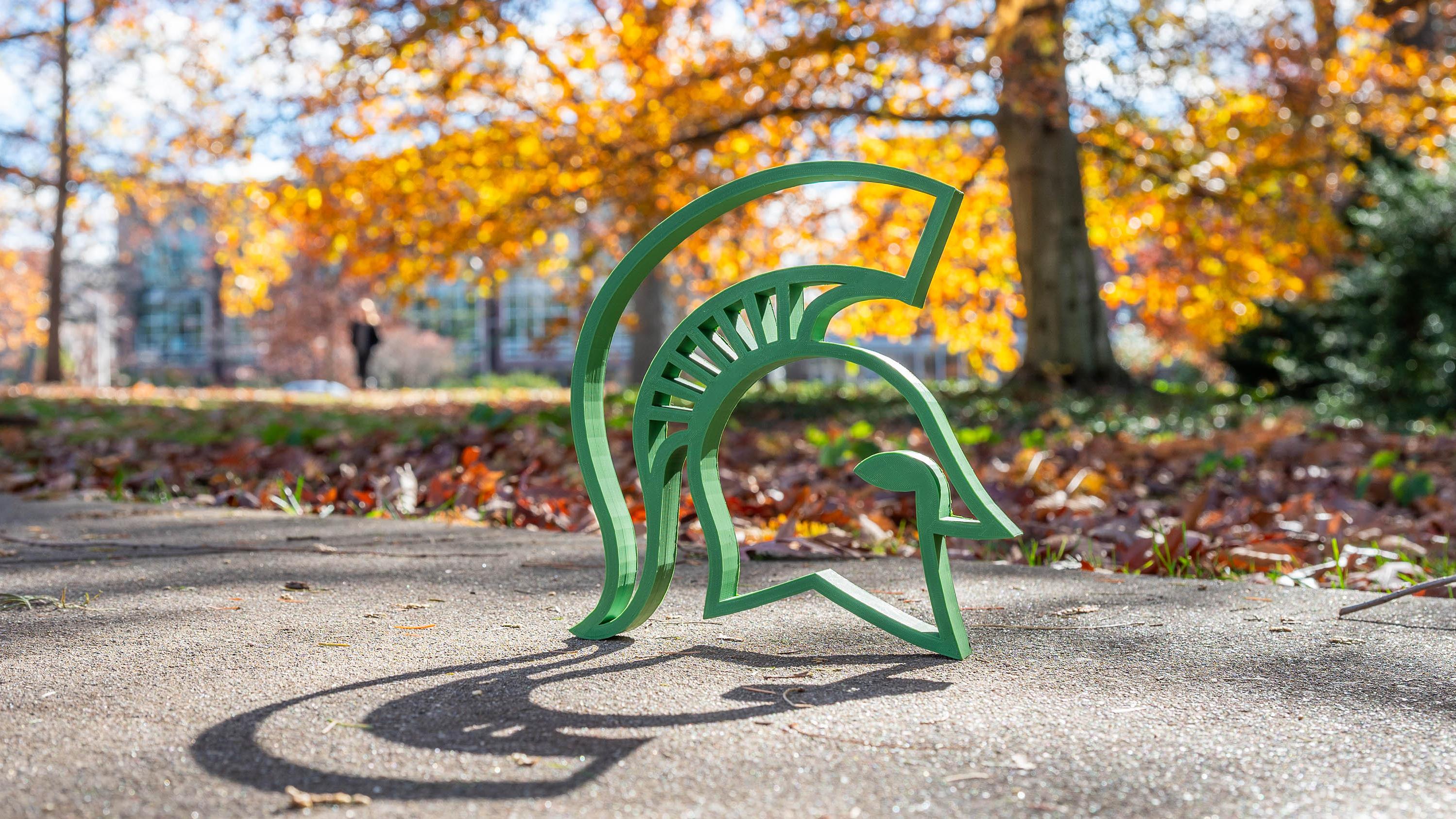 3D printed green Spartan helmet sculpture on a sidewalk with colorful autumn trees and fallen leaves in the background.